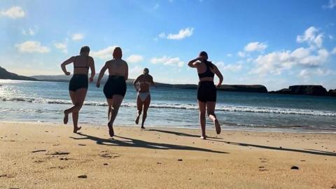 Four swimmers run across the beach towards the sea with bright sunny blue sky above
