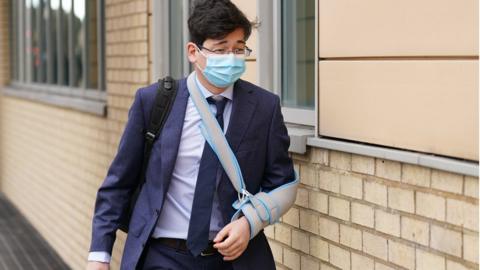 Jonathon Dean arrives at Cambridge crown court. He is wearing a surgical mask, a blue suit and has his left arm in a sling.