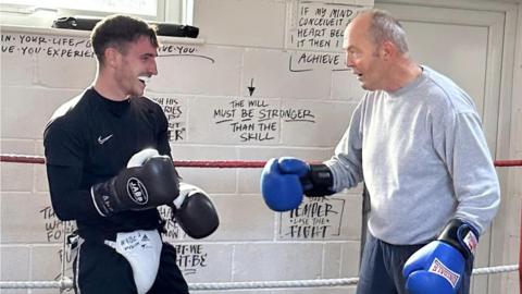 Two men in boxing gloves practising, the younger on the left, an older man on the right.