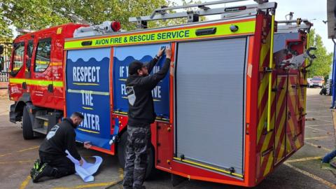 Two men putting the new wrap onto a fire engine. Their backs are to the camera. They are wearing black hoodies with a Shades Wraps logo on the back. Both panels are royal blue with white capital letters which are underlined in yellow. The first panel reads 'Respect the water' and the second reads 'If you fall in, float to live'.