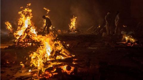 Workers and relatives stand around burning funeral pyres at night