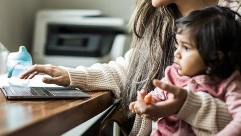 Stock image of a mum holding an infant while working on a laptop.