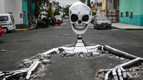 A huge cardboard cardboard skeleton emerges from a street in Mexico City