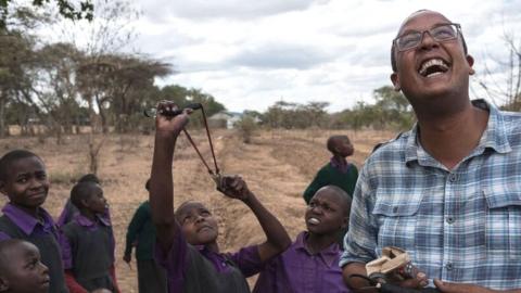 A Kenyan man laughs as children aim a catapult at the sky