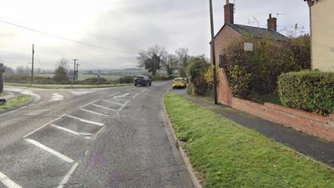Rural road on the edge of a village with houses to the right and fields to the left