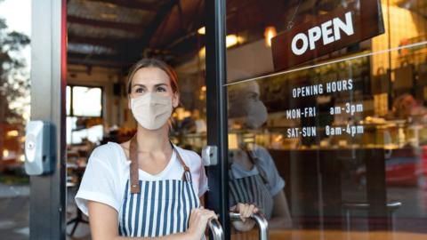 A cafe owner wearing a mask looks out of the front door or her cafe