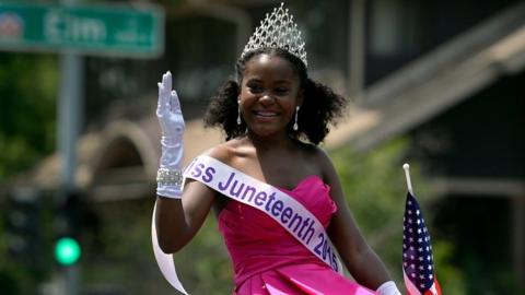 Young woman with Miss Juneteenth sash in pink dress waves at crowds