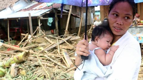 A woman stands with her baby in front of damaged houses and fallen trees in Barangay San Mateo Borongan, eastern Samar, on 17 December 2017, after Tropical Srtorm Kai-Tak blew through the area