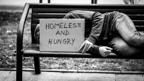 A homeless person lying on a bench with a sign that says homeless and hungry