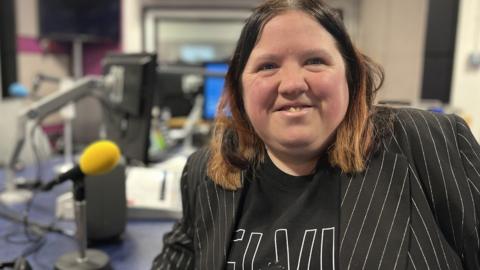 A woman with dark hair and a suit jacket sits in a radio studio