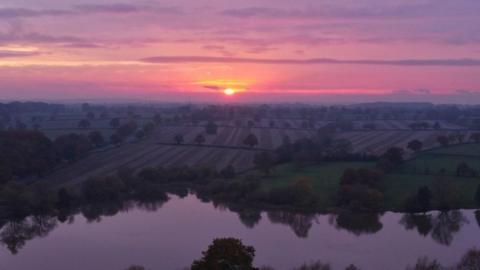 The sun is setting in the middle of the horizon with a sky filled with pink, purple and yellow hues. There is a lake and fields with trees in the foreground.