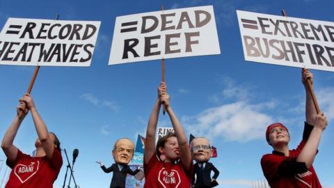 Young environment activists hold protest signs up in front of comedians dressed as Labor leader Bill Shorten and Prime Minister Scott Morrison on 5 May 2019 in Canberra, Australia