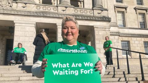 Colleen McCallion standing in front of Stormont holding a sign reading "What are we waiting for?"
