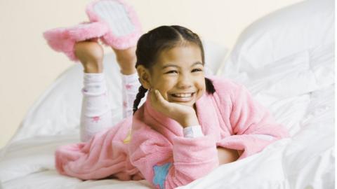 Girl lying on her front on a bed wearing pyjamas, dressing gown and slippers