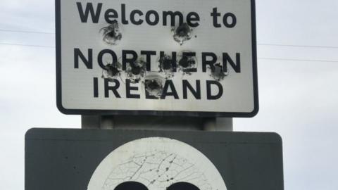 bullet holes riddle the Welcome to NI sign