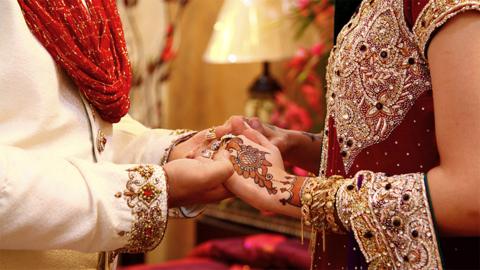 Bride and groom are holding their hands of each other. The groom is wearing a cream white outfit with a sparkly red and the bride is wearing a maroon outfit with intricate silver and golden designs, with orange and red coloured henna on the front of her hands.