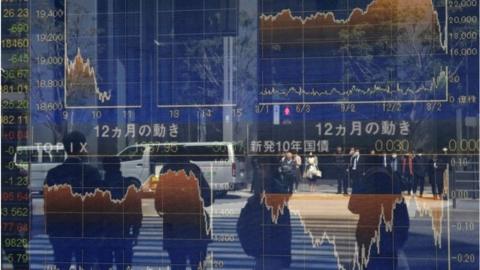 Pedestrians reflected in a window stand in front of a quotation board displaying the numbers on the Tokyo Stock Exchange in Tokyo.