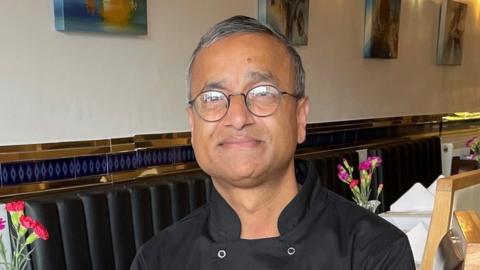 Karim Ullah is sat on a chair in his restaurant. He is an Asian man and has short dark hair that's going grey and is wearing round spectacles. He is wearing a black chef's overall and behind him you can see tables in his restaurant with purple flowers on them. There are also a few bits of artwork on the cream walls behind him.
