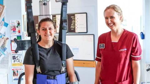 Megan is on the left being held up standing using hoist straps and smiling. Elly is on the right in a red uniform smiling at the camera