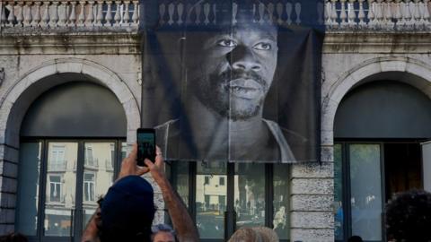 A demonstrator takes a picture of the large portrait of Bruno Candé