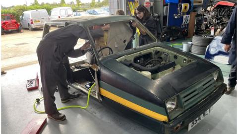 Two students in dark overalls working on the shell of an old car