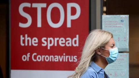 Woman wearing facemask walking by a sign reading: Stop the spread of Coronavirus