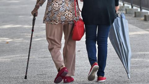 A rear view of a woman with a walking stick walking with a carer in a Singapore park