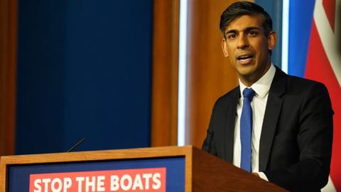 Rishi Sunak at a press conference, standing in front of a sign that says "Stop the Boats"