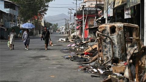People walk past a burnt vehicle and rubble on a street in Churachandpur in violence hit areas of northeastern Indian state of Manipur on May 9, 2023.