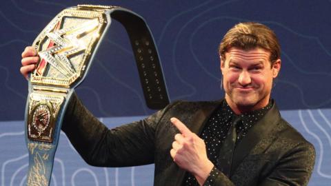 A man with tied back blonde-brown hair stands in front of a giant screen displaying a dark blue/light blue swirl pattern. He's dressed smartly in a black suit and tie, with a black shirt with white polka dots underneath. He's pointing towards a wrestling championship belt that he's holding up at shoulder hight. It's gold, with a WW logo on it. The design is encrusted with diamond-style gems.
