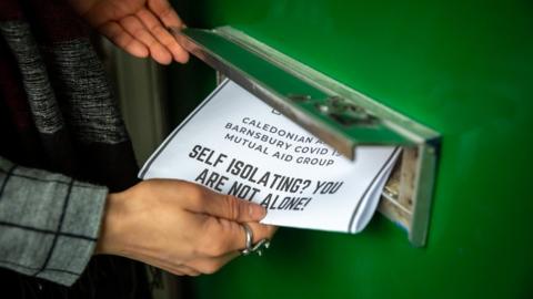 A leaflet being pushed into a letterbox reads "self-isolating - you are not alone"