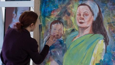 A woman paints a large picture of a woman holding a baby