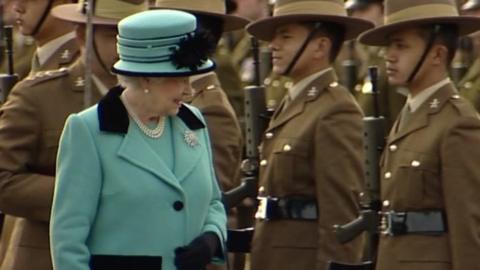 The Queen in a blue dress suit with Gurkhas behind her