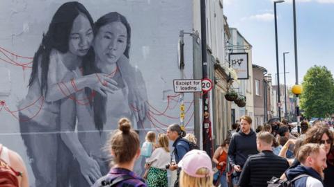 A black and white piece of street art of a mother and a daughter playing with red string. Crowds of people are filling the street