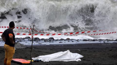 A policeman stands next to the body of a body-boarder killed by a shark, covered by a white cloth next to his body-board on 21 February 2017 on a beach in Saint-Andre, on the French island of Reunion in the Indian Ocean