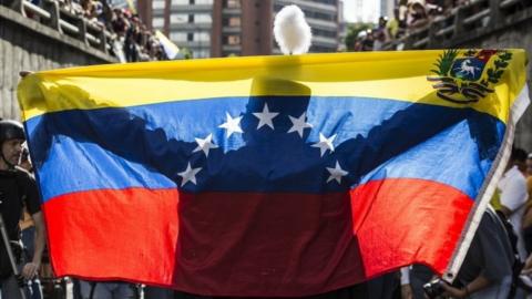 person holds a Venezuelan national flag before dozens of demonstrators during an anti-government rally in Caracas, Venezuela, 24 July 2017