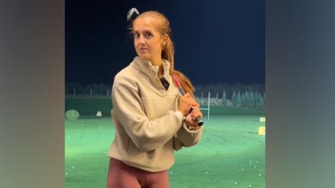 A young woman in a cream-coloured fleece and lilac leggings stands in front of a grass-covered driving range with a golf club resting on her shoulder. Her eyebrows are raised and she looks slightly shocked with a hint of amusement, like she can't quite believe what she just heard.