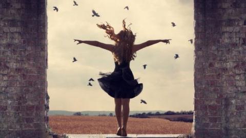 A woman looking out at a view with her arms in the air with birds in the sky