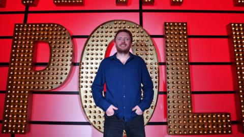 Chris McCausland standing in front of the Live at the Apollo sign.