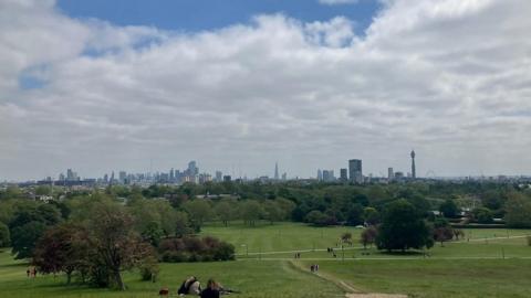 A view down Primrose Hill, London, with people sitting on the grass near trees and the city skyline on the horizon. Above the sky is largely filled with white clouds.