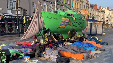Protesters in sleeping bags surround green boat in the middle of Castle Street
