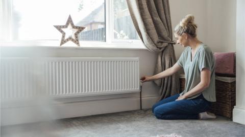 Side view of a young woman kneeling on the living room floor, adjusting the thermostat on the radiator.