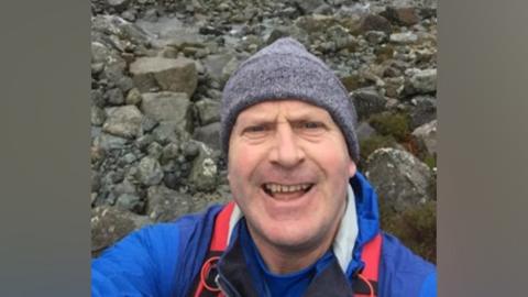 Neil McGibbon taking a selfie, wearing a grey hat on a rocky landscape