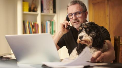 A man is on the phone, petting his dog while also surveying assorted papers scattered around his open laptop