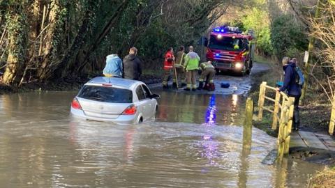 Car in flood water with two people sitting on its roof with firefighters nearby