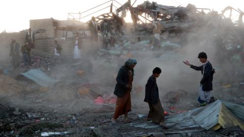 people walking in front of smoke and ruined houses in the aftermath of an airstrike in Saada