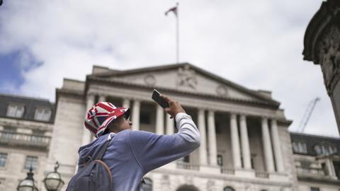 A general view of the Bank of England in London on 3 August, with a person in the foreground taking a picture on mobile phone