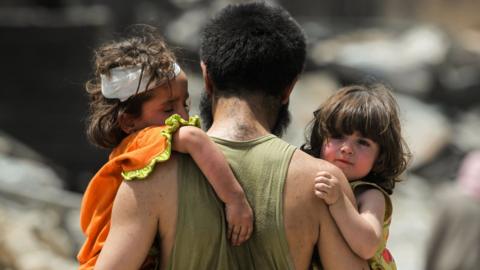 A man carries two young children as they flee fighting in the Old City of Mosul, Iraq (2 July 2017)