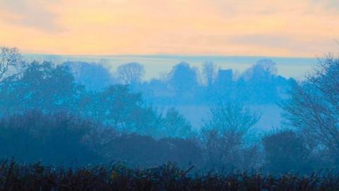 The fog has created layers of light which look like blue horizontal stripes sandwiched between the dark foreground and a pale orange sky.