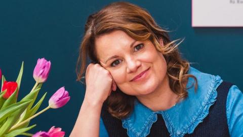 Louise Taylor resting the side of her head on a fist smiling at the camera wearing a blue large collard blouse over under a dark top with red and purple tulips in the bottom left